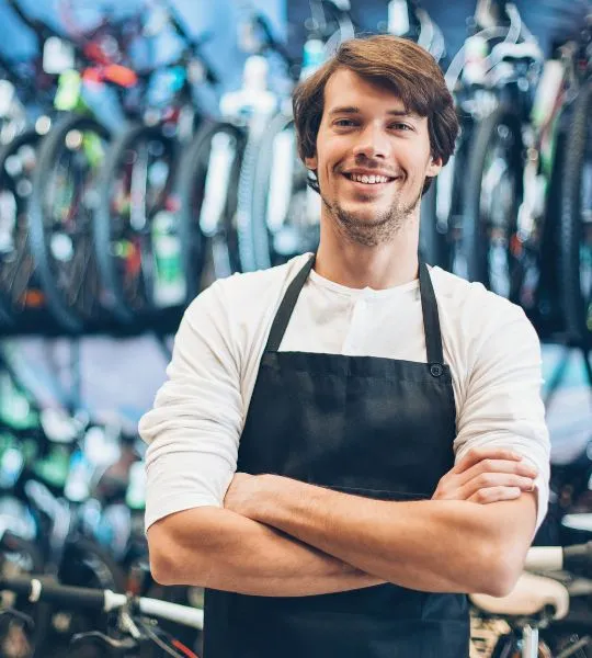 Bike shop owner in a black apron standing with arms crossed in front of bicycles.
