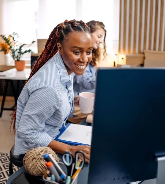 Two coworkers at a desk, one holding a mug while looking at a computer.